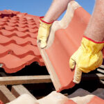 Roof repairs, worker with yellow gloves replacing red tiles or shingles on house with blue sky as background and copy space.